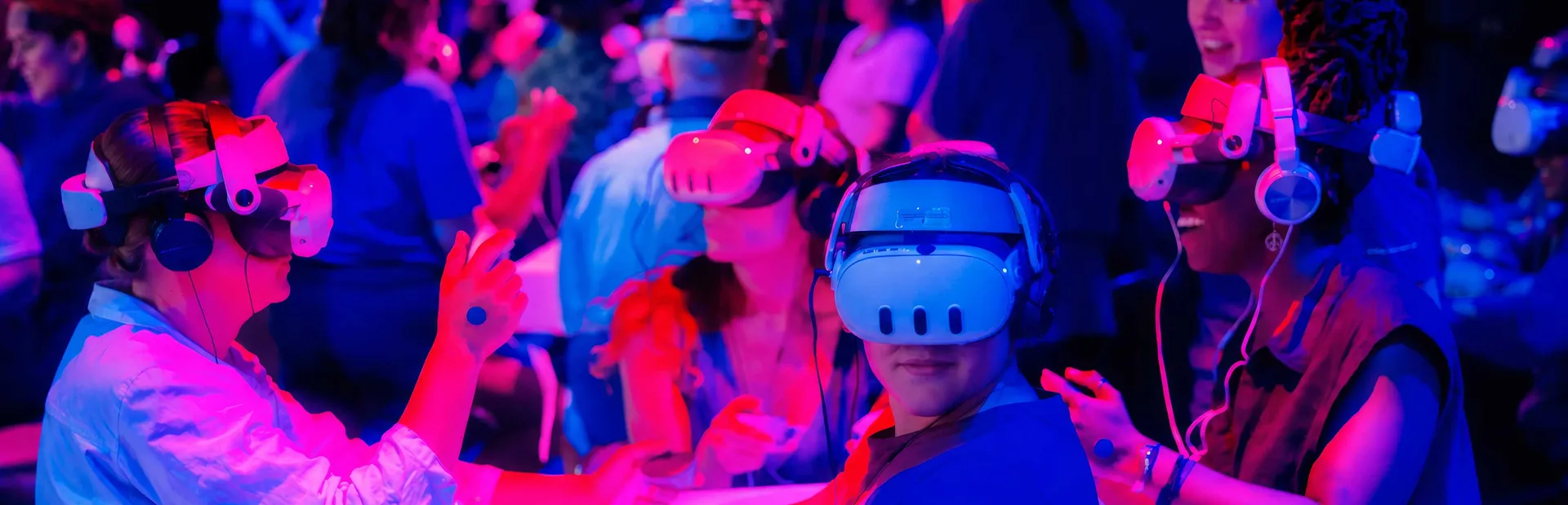 The Sum of Us - visitors wearing VR headsets and headphones reaching out with their hands during The Dream Machine Experience, lit in red and blue