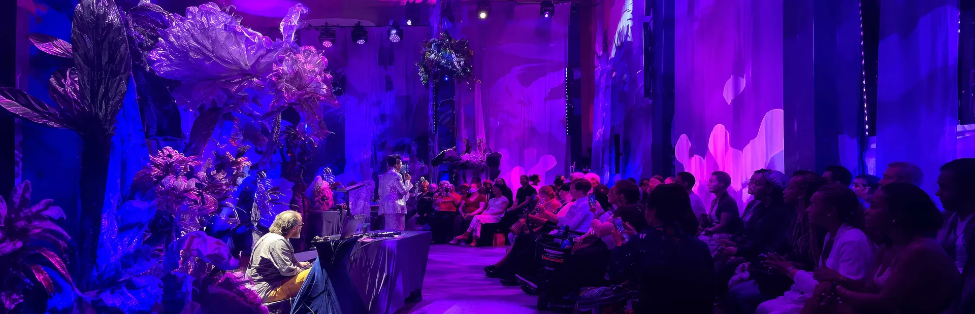 The Sum of Us - Nona Hendryx in white singing into a microphone on a purple-lit stage dressed with translucent sculptural flowers, seated audience alongside